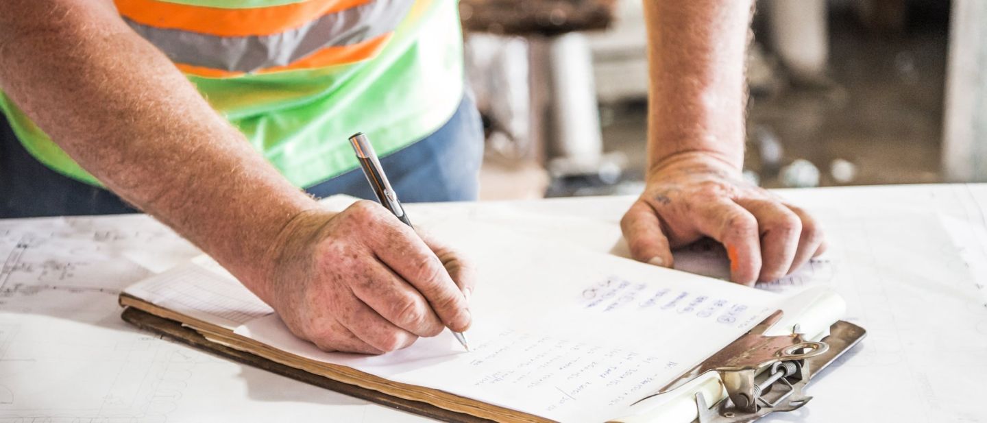 Hände notieren auf einem Klemmbrett über ausgebreiteten Bauplänen auf der Baustelle